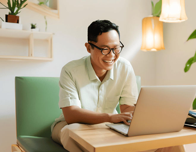 A brightly lit room features an Indonesian man with a relaxed posture, focused on his laptop, evident joy visible on his face.
